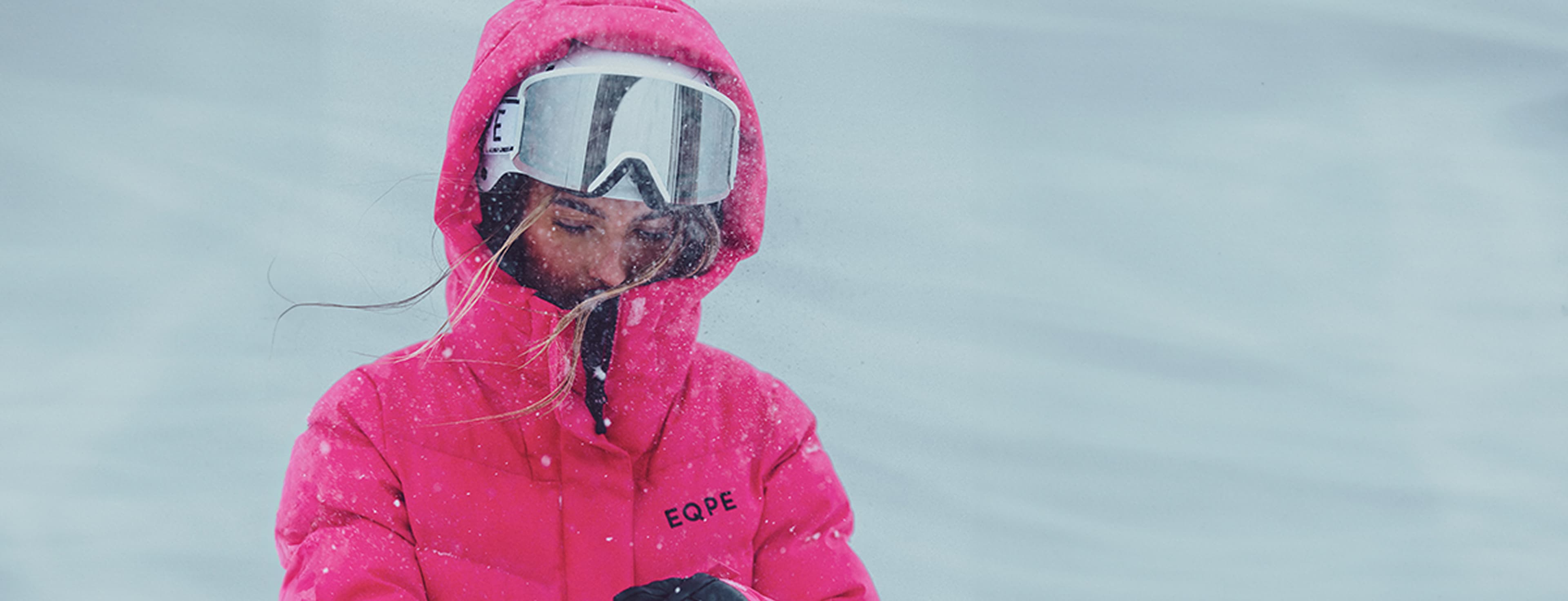 Person in pink jacket and goggles standing in falling snow, looking down with hair blowing in the wind.