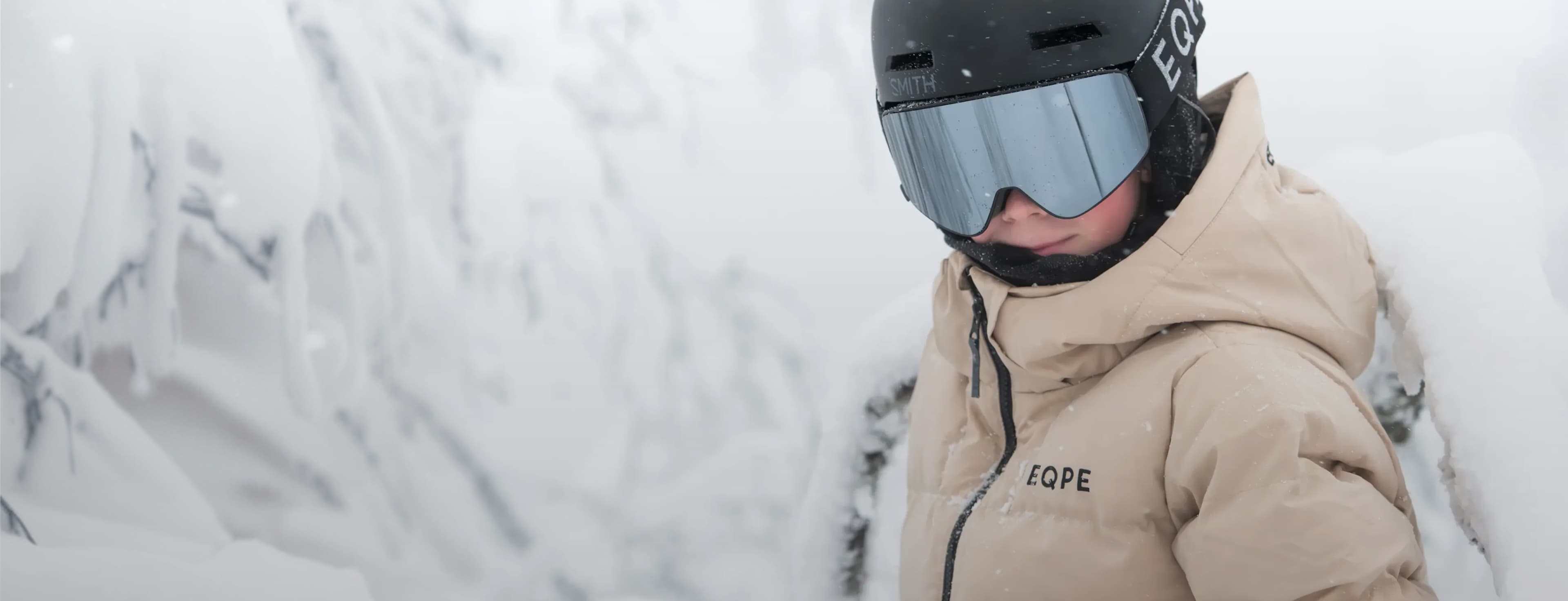 Person in a beige winter coat and black helmet with goggles stands in a snowy landscape, surrounded by frosted trees.