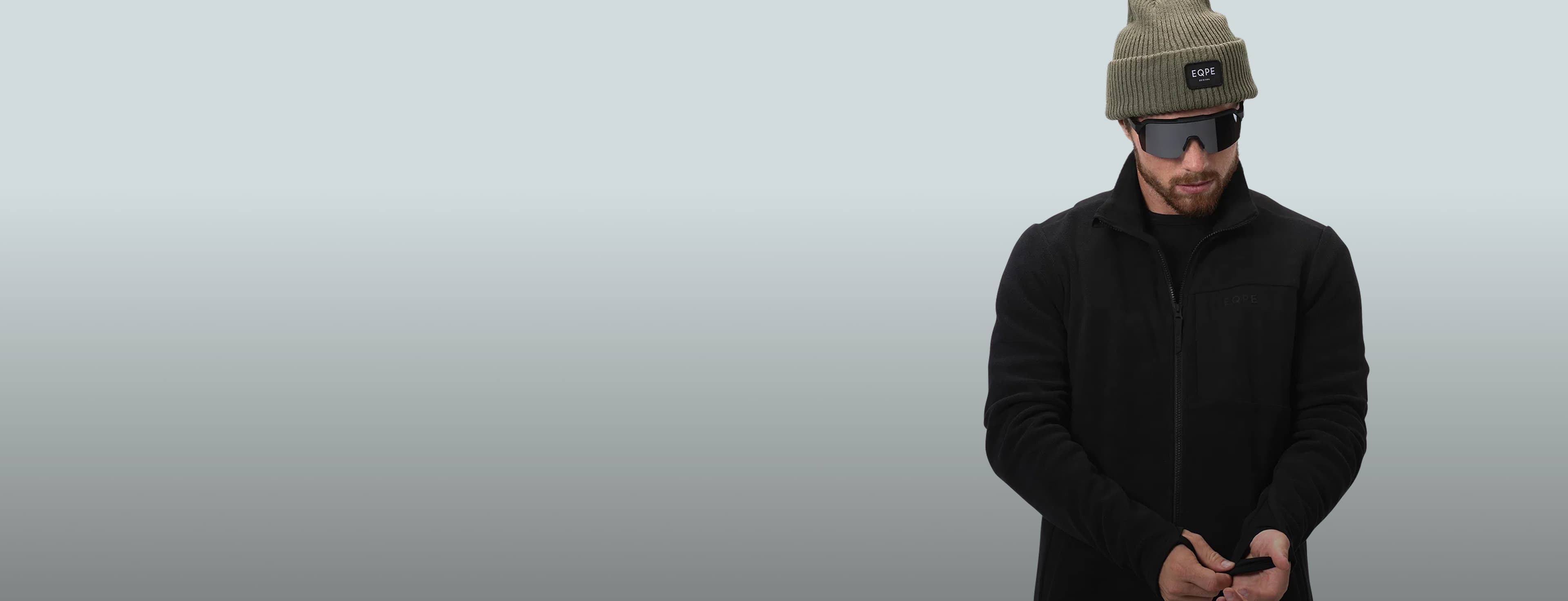 Man in black jacket and beanie, wearing sunglasses, looking down.