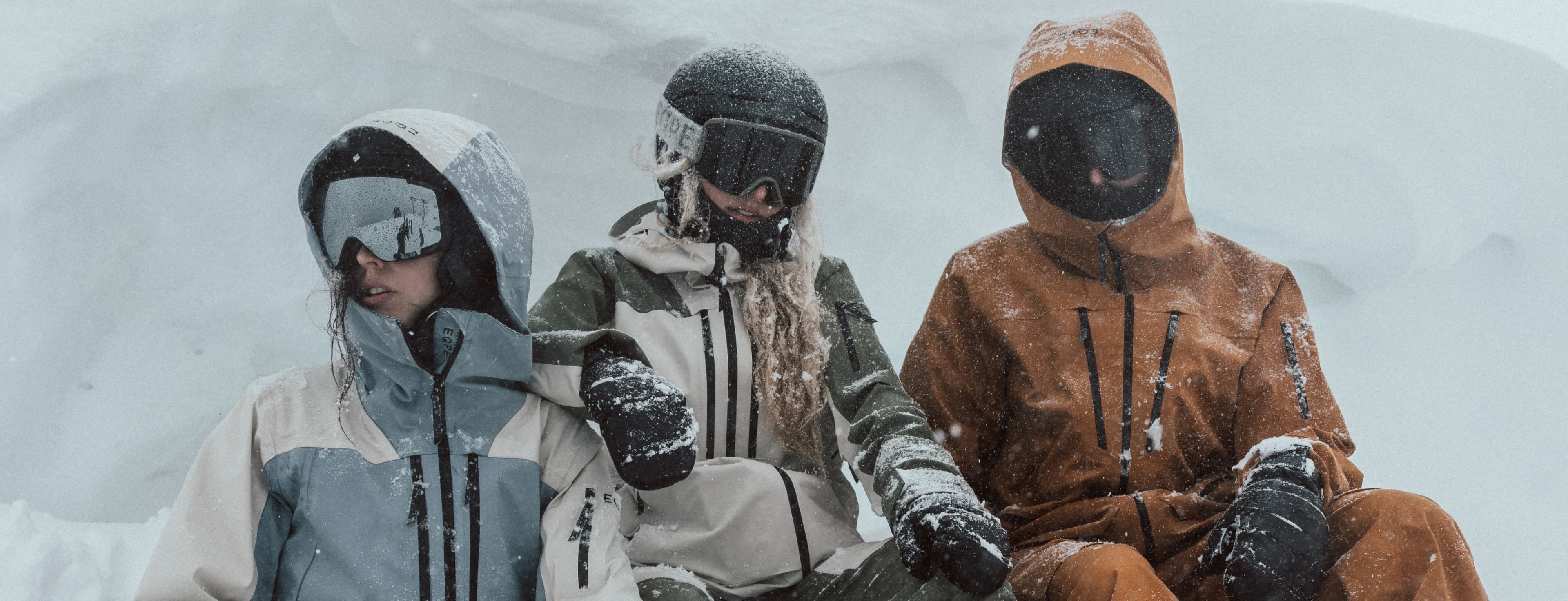 Three people in ski wear, sit in the snow during a snowfall.