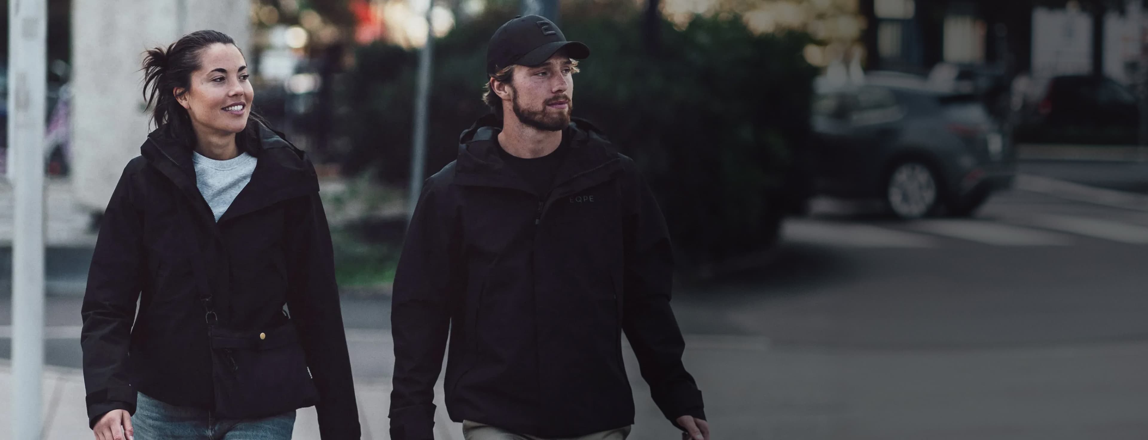 A man and a woman in black jackets walk confidently on a city street, smiling, with blurred urban background and cars passing by.