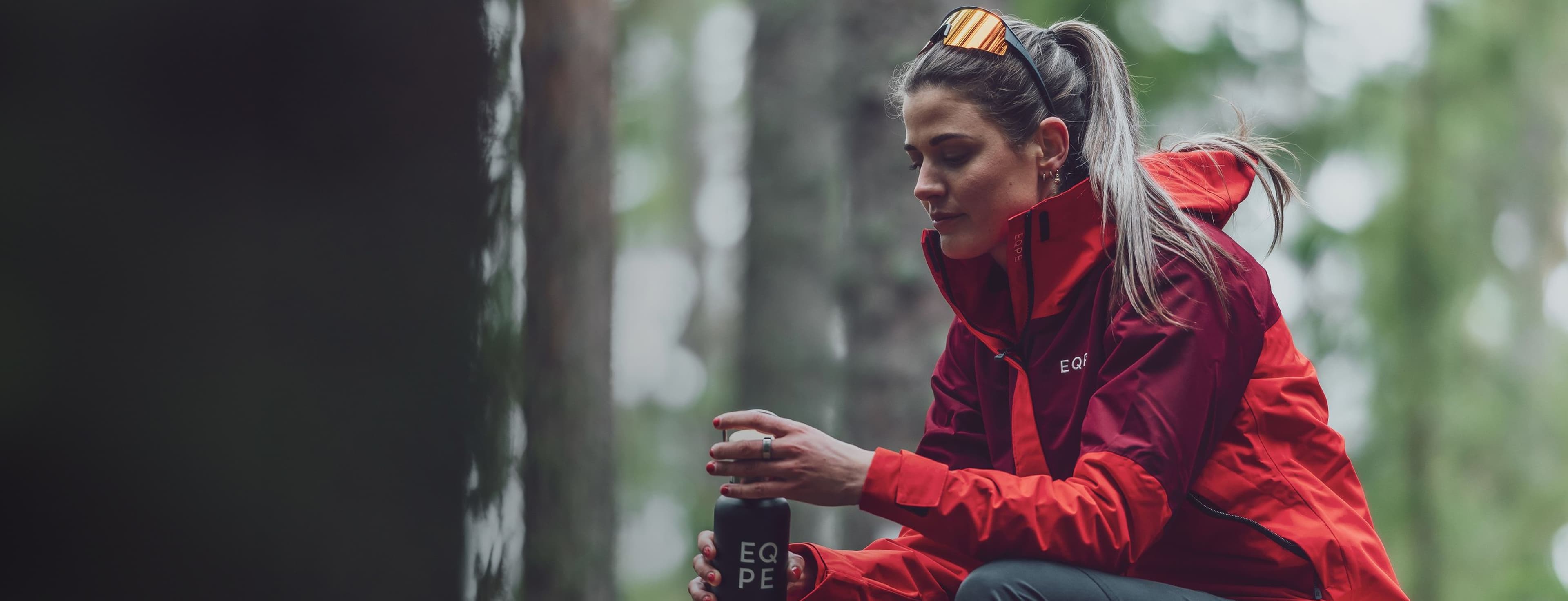Woman wearing a red jacket, seated on a mossy rock in a forest, holding a black water bottle.