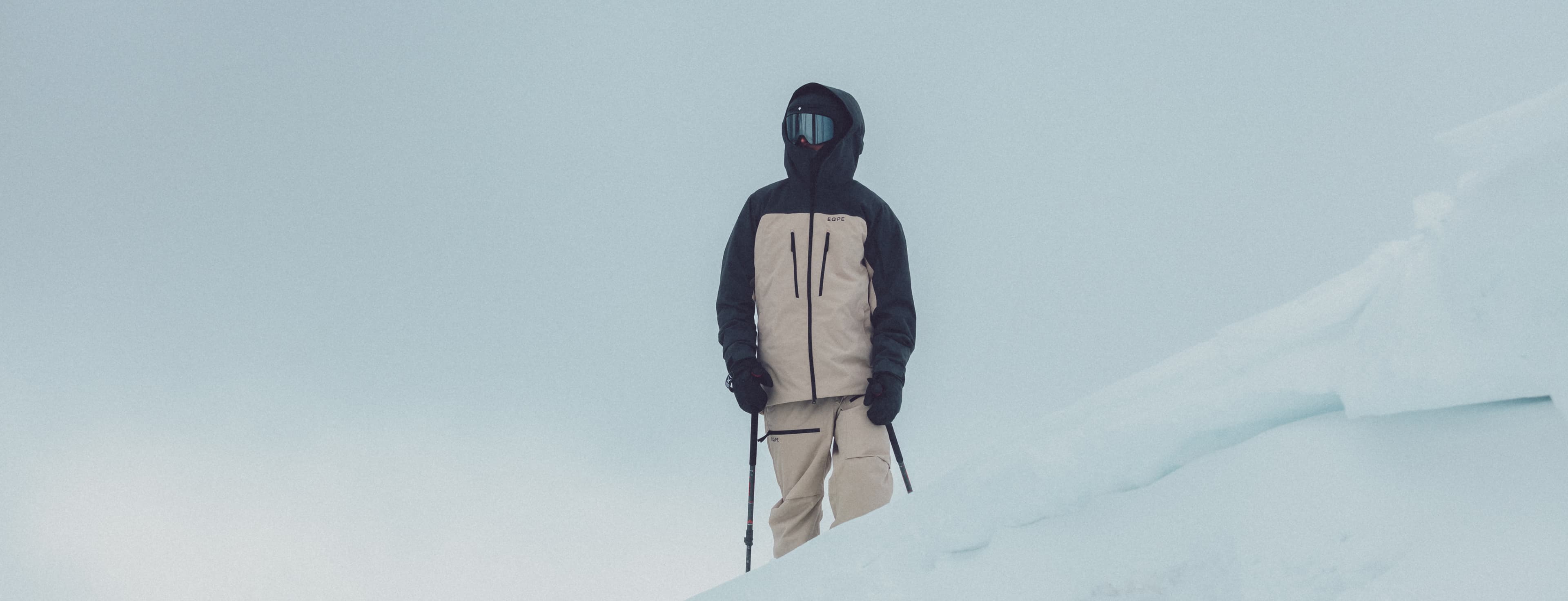 Person in winter gear stands on a snowy slope, holding ski poles, against a cloudy sky.