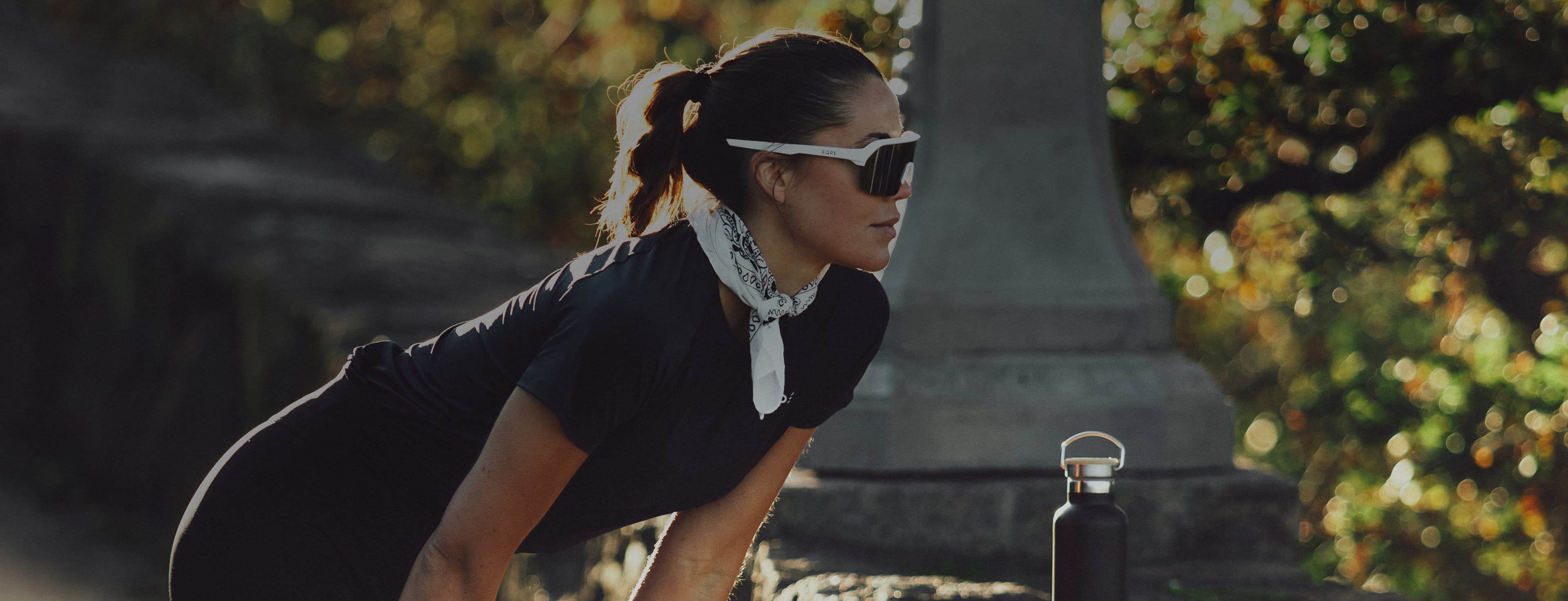 Woman in activewear leaning on a rock, wearing sunglasses and a white scarf, with a water bottle nearby, outdoors.