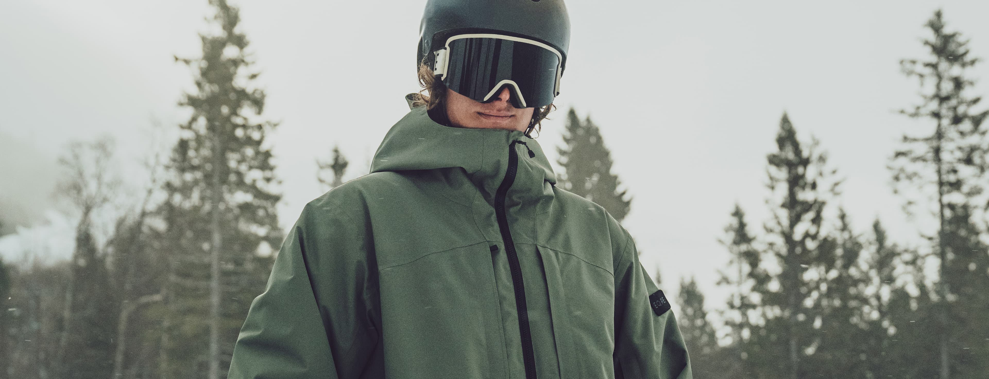 Person in a green ski jacket, standing outdoors with snow-covered trees in the background.