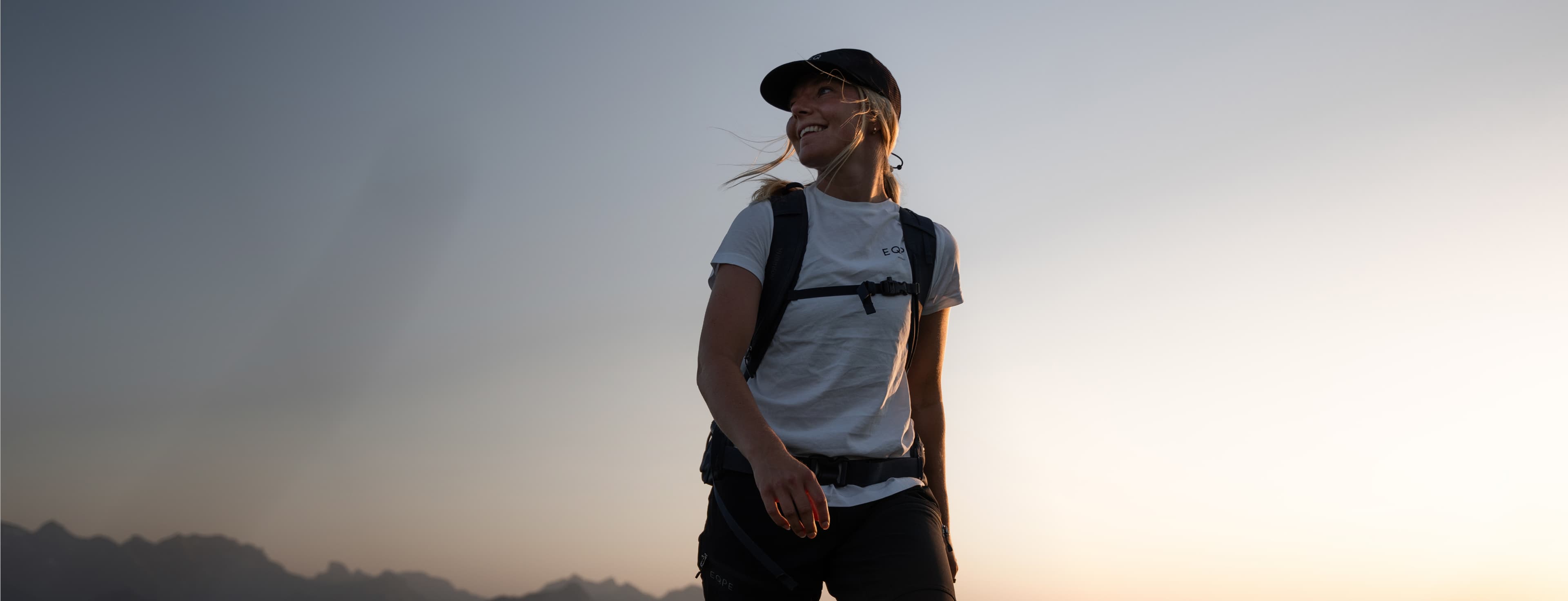 A person wearing a cap and backpack stands in front of a dusky sky, smiling and looking to the side, with mountain silhouettes in the background.