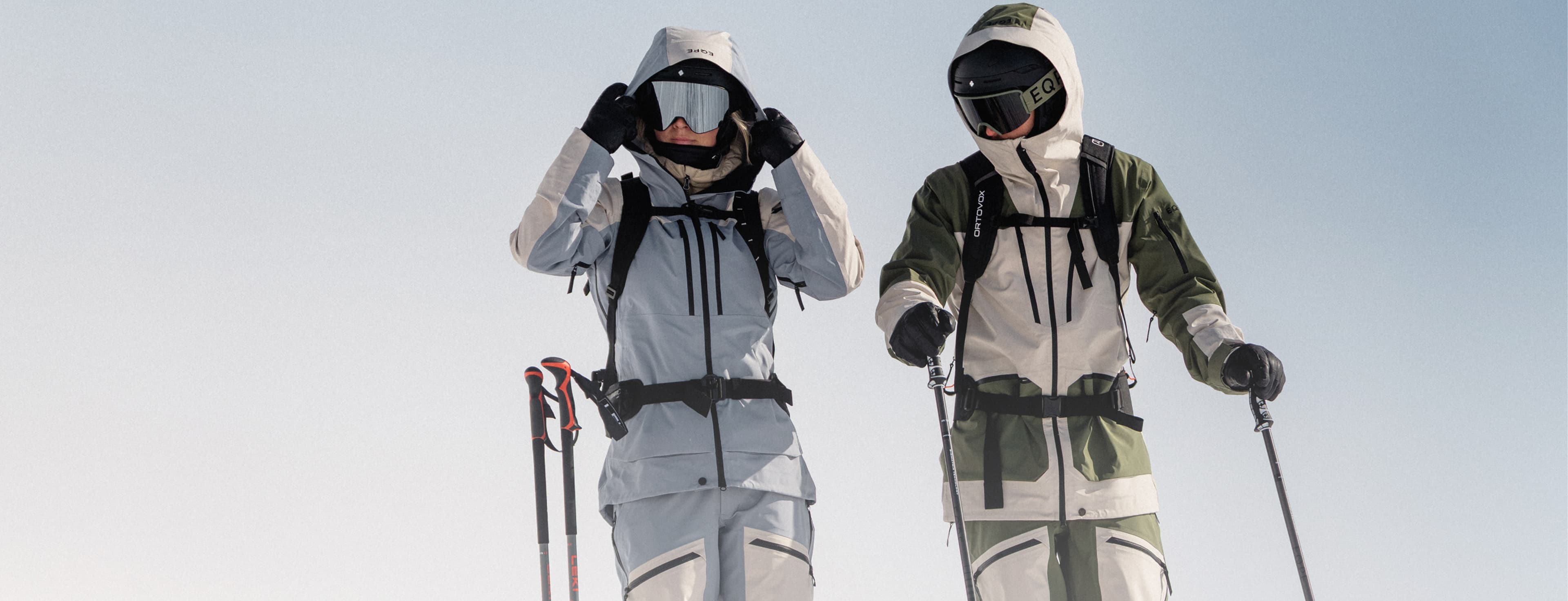 Two skiers in heavy winter gear and helmets, standing with ski poles against a clear sky background.