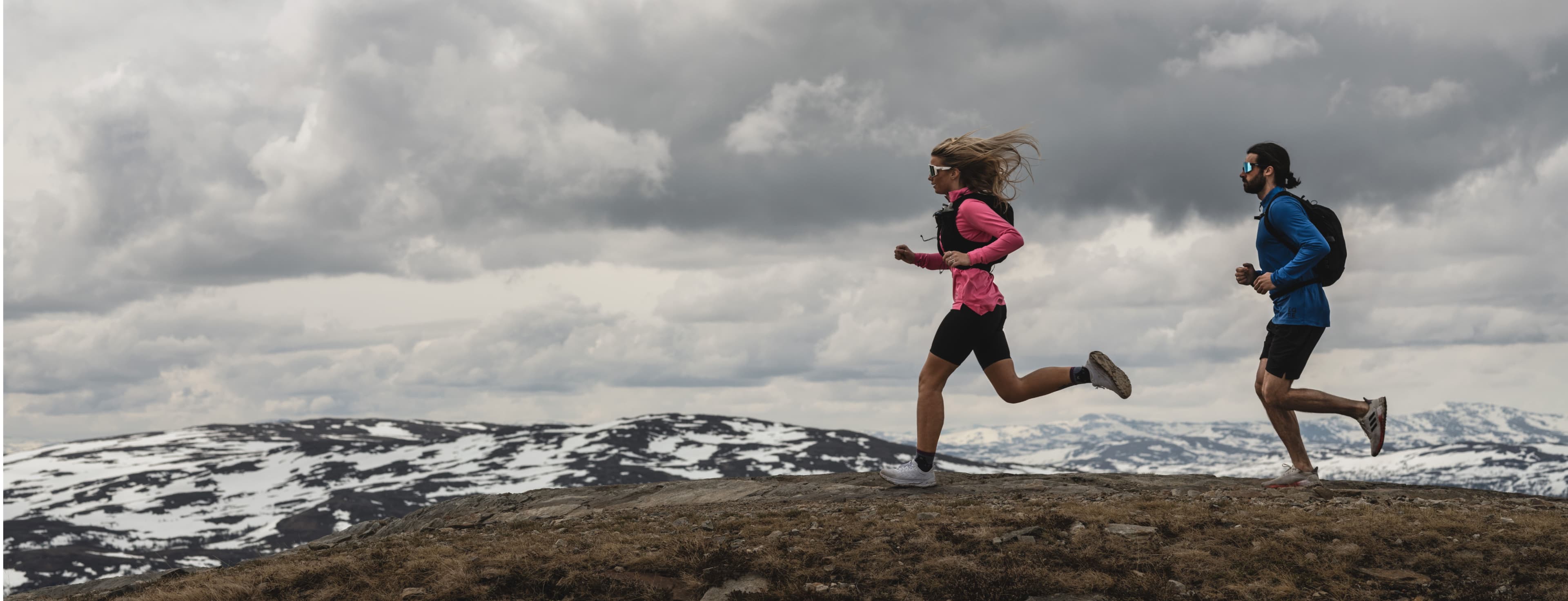 Two people running on a mountain trail, wearing athletic gear, under a cloudy sky with snow-dusted peaks in the background.