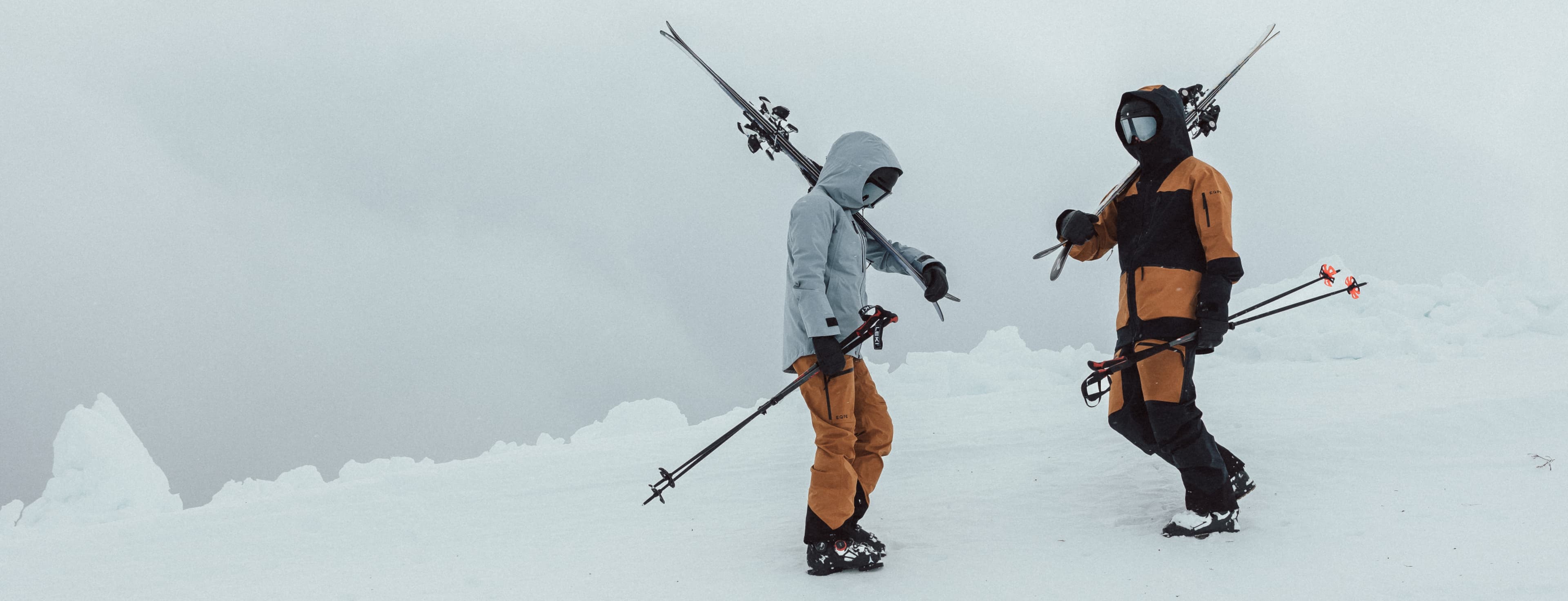 Two people in ski gear stand on a snowy mountain slope under a cloudy sky.