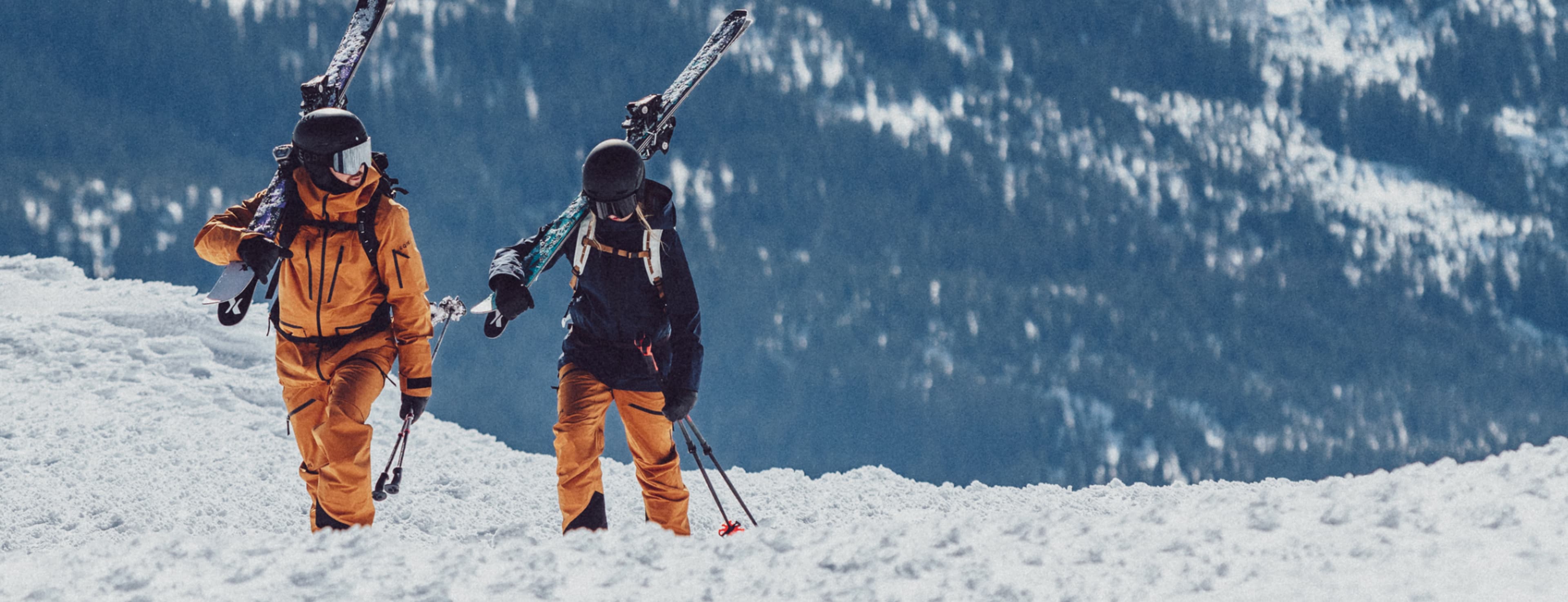 Two skiers carry skis on their shoulders, walking through snowy terrain with a forested mountain backdrop.