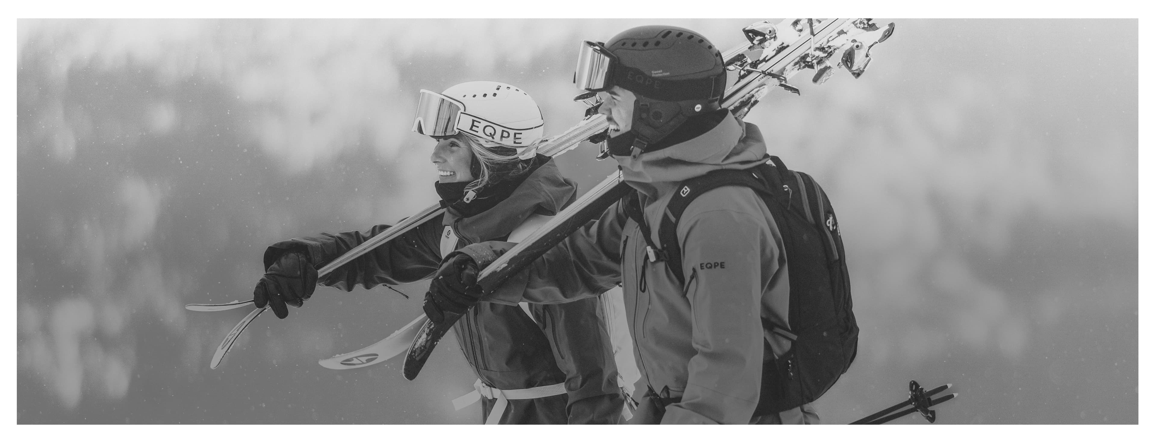 Two skiers in gear walk side by side, carrying skis over their shoulders, against a snowy mountain backdrop. Black and white image.