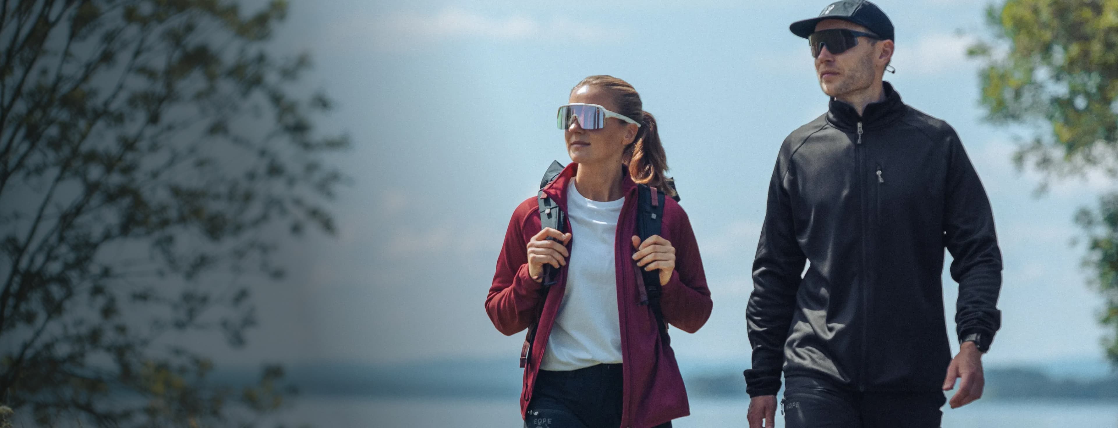 Two people in sunglasses and outdoor gear walk by a lake, surrounded by trees, on a sunny day.
