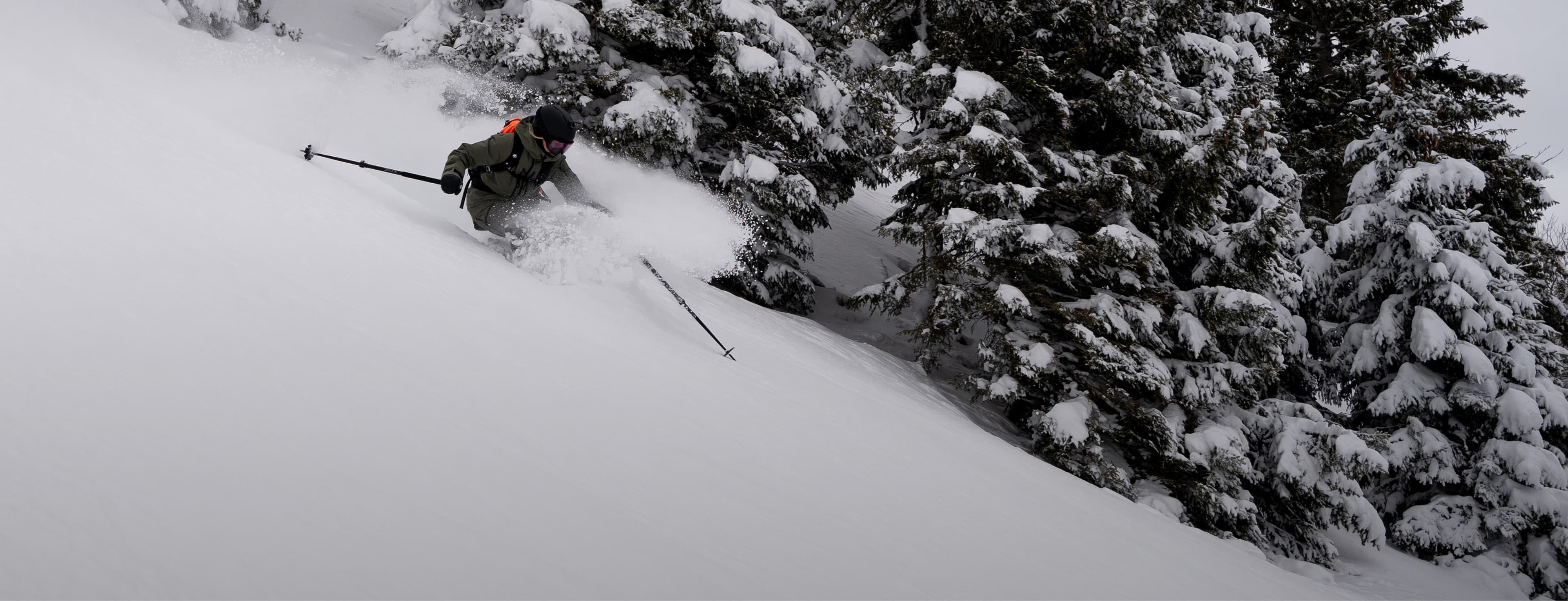 A skier navigates through fresh snow on a steep, tree-lined slope under a cloudy sky.
