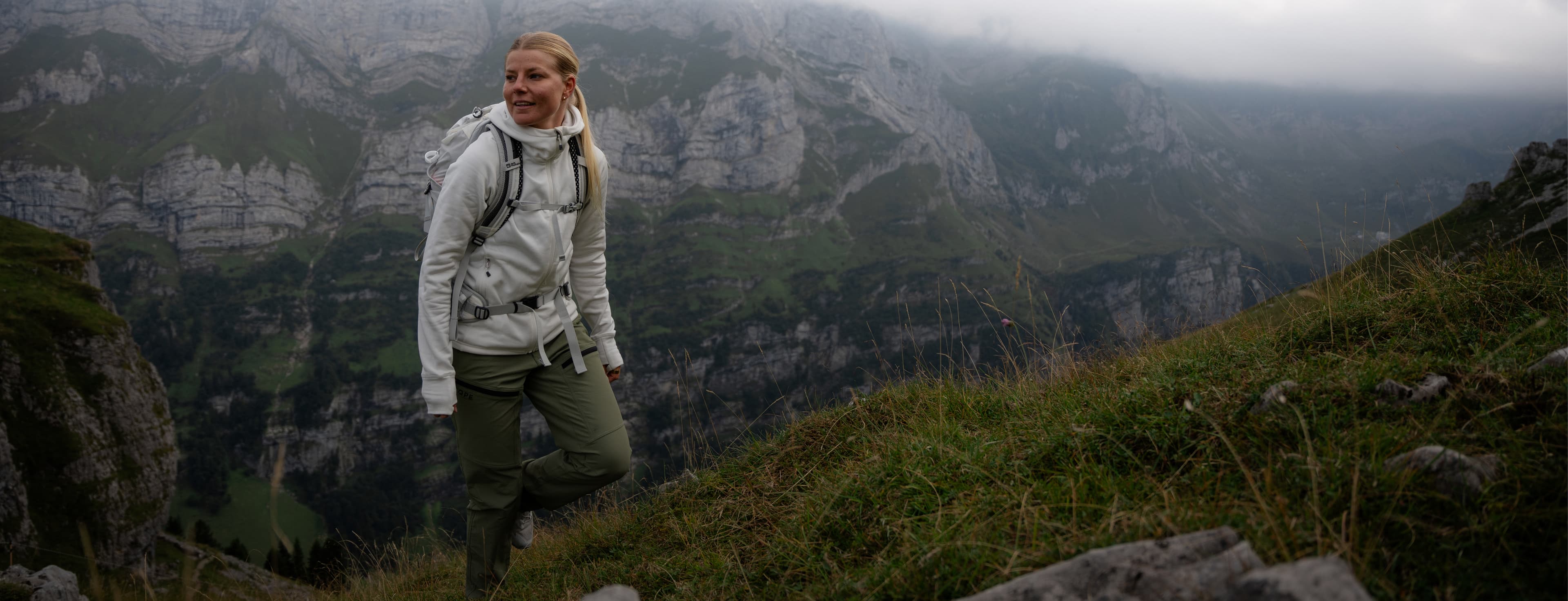 A person in hiking gear stands on a grassy slope, with misty mountains in the background under a cloudy sky.