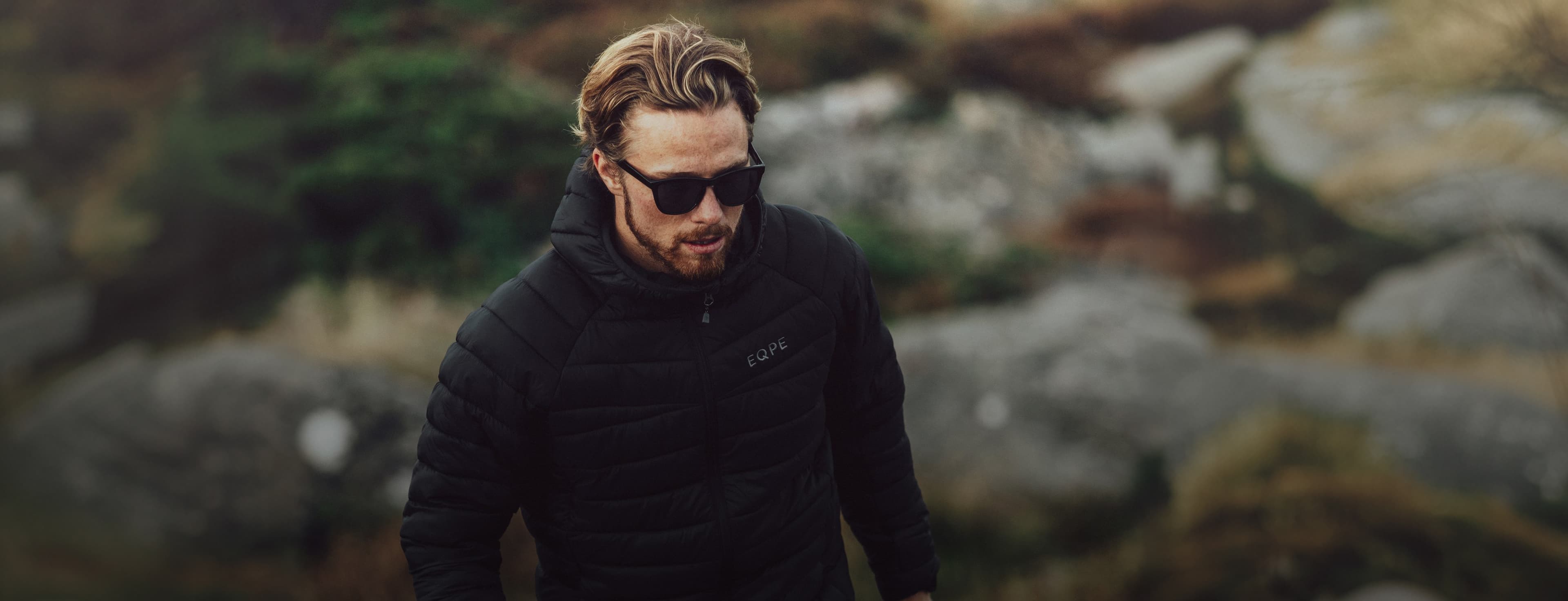 Man in sunglasses and a black puffer jacket walking outdoors on a rocky terrain with blurred greenery in the background.