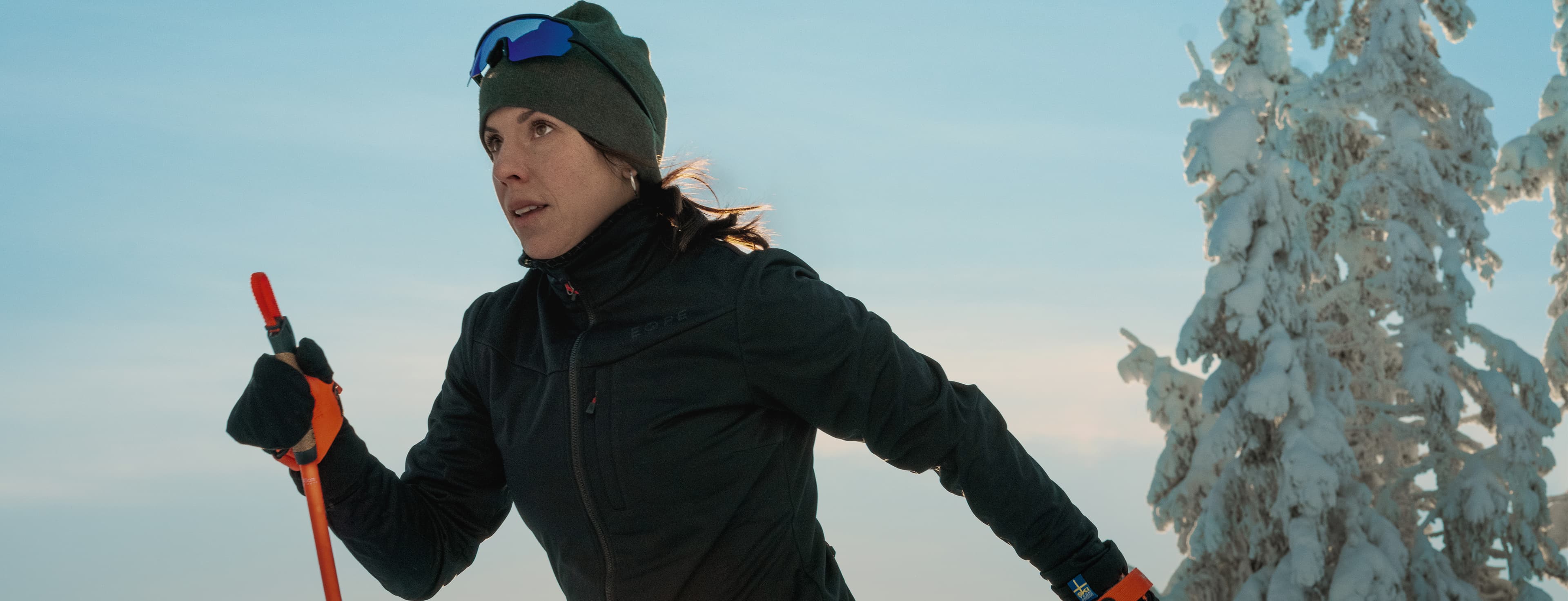Woman cross-country skiing in winter, wearing a black jacket and hat, holding ski poles, with snowy trees and a clear sky in the background.