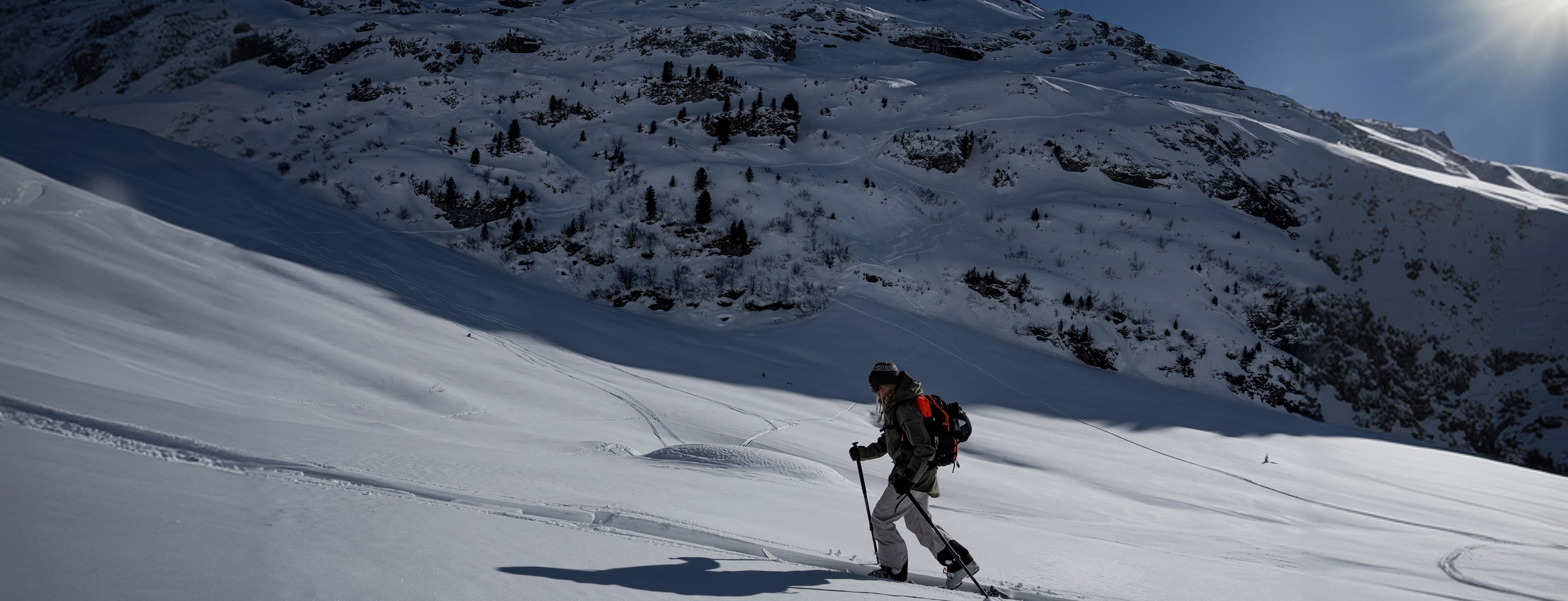 A skier in a black jacket and red backpack ascends a snowy mountain under a clear blue sky with the sun shining brightly.