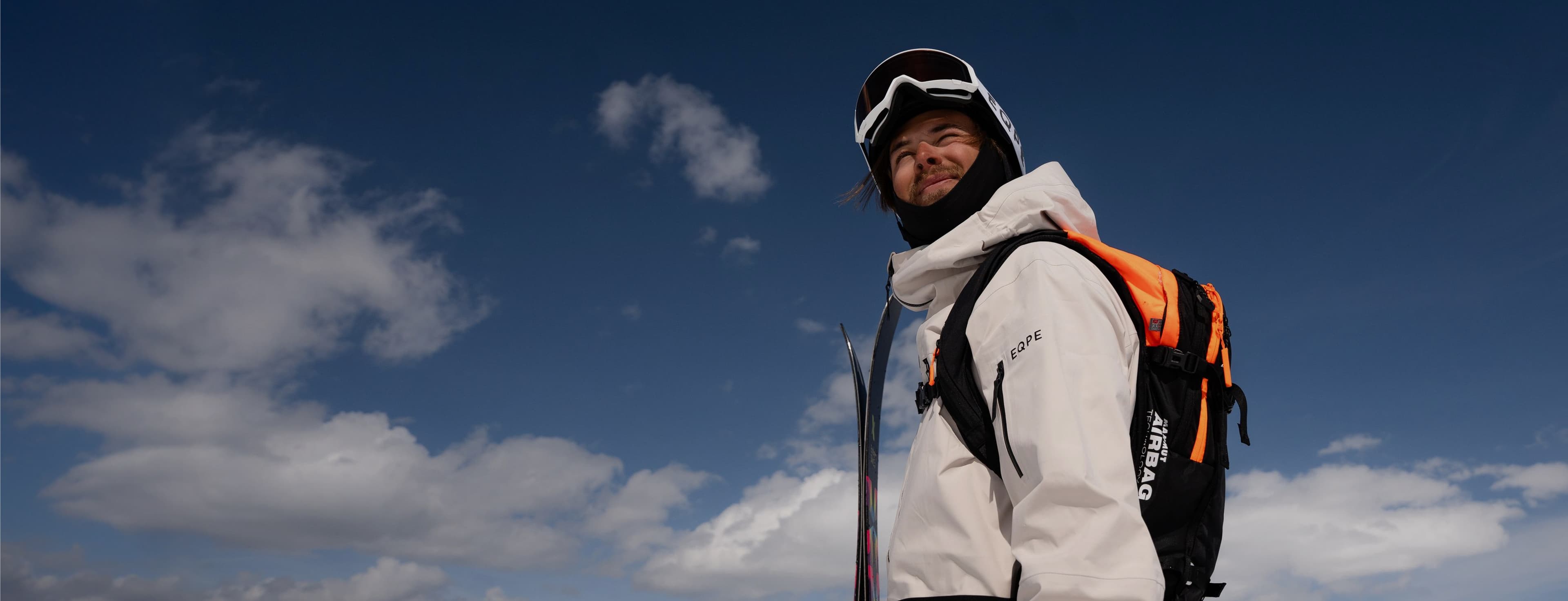 Person in beige winter gear with ski goggles and backpack gazes upward against a blue sky with scattered clouds.