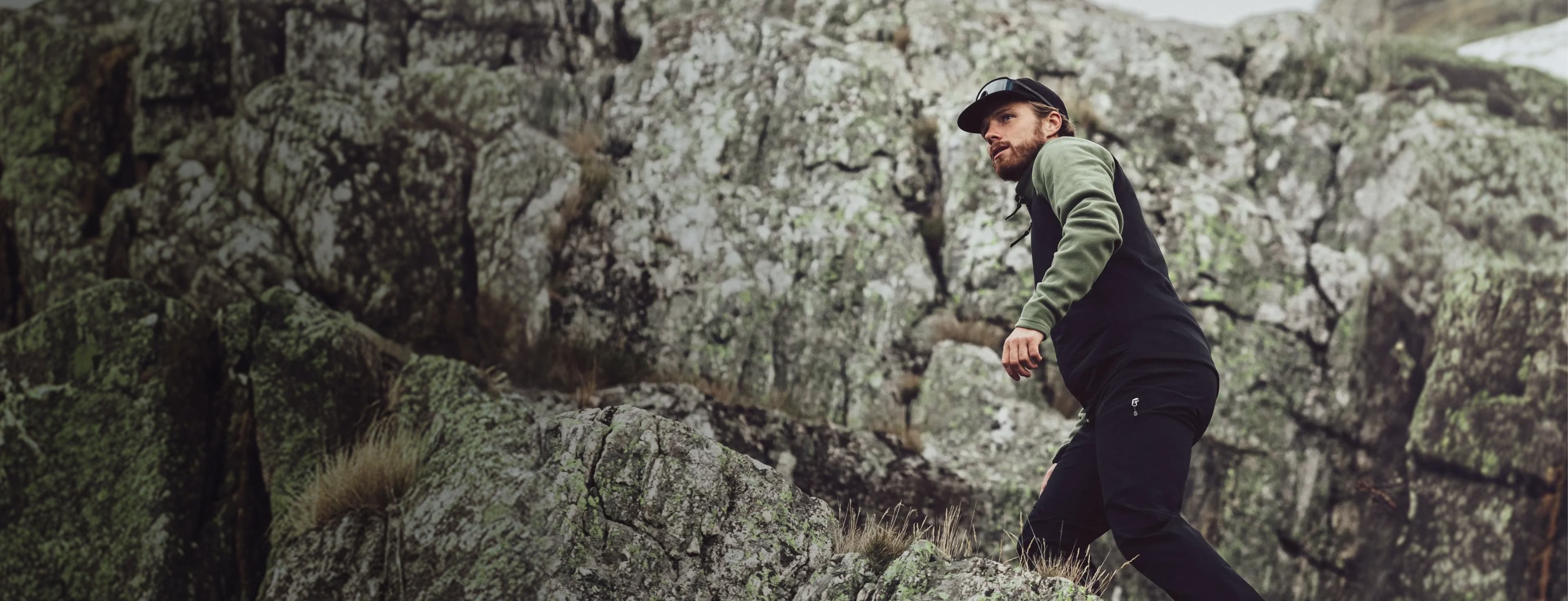 Man wearing a cap and hiking clothes climbing rocky terrain, looking determined against a rugged rock background.