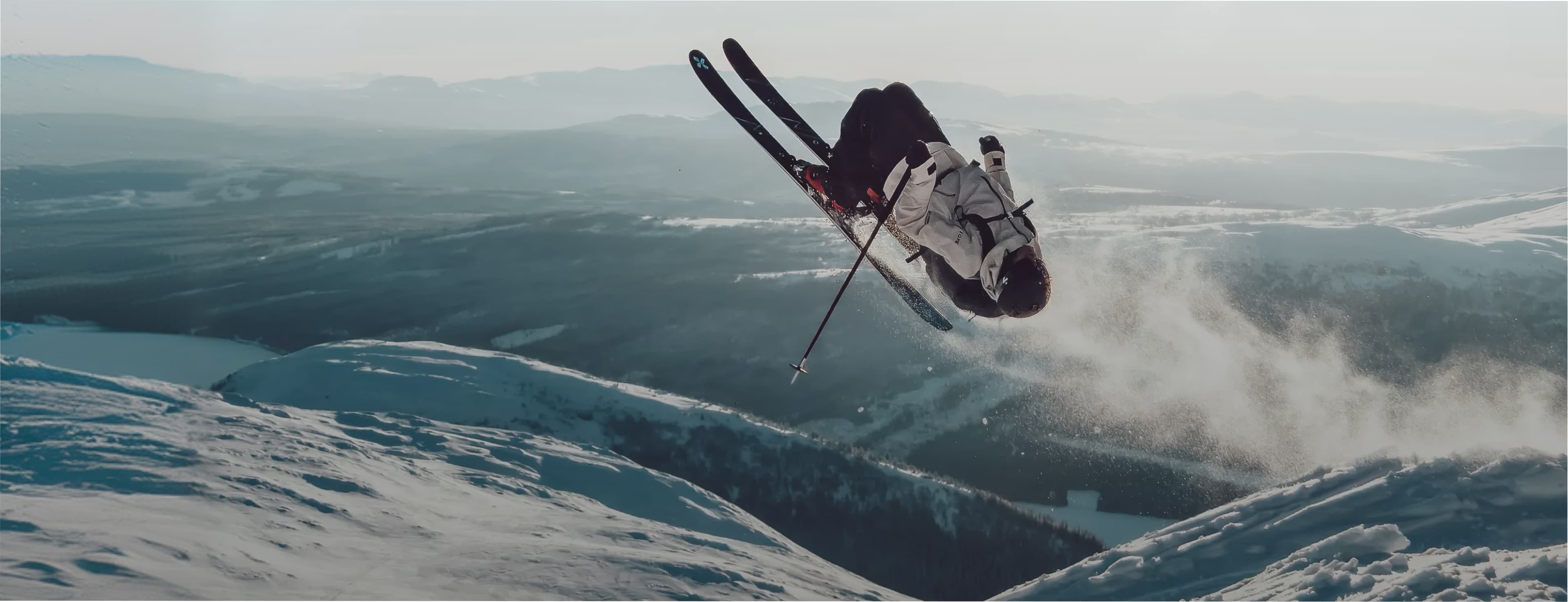 A skier performs a mid-air flip against a backdrop of snowy mountains and expansive landscape under a cloudy sky.