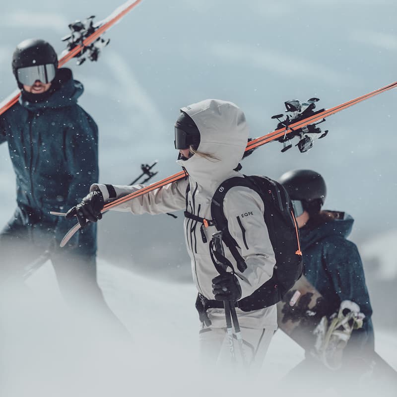 Three skiers in winter gear carrying skis on their shoulders, walking through snow on a mountain with a foggy background.